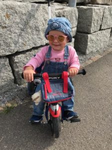 Aileen, 2 Jahre, ist ein aufgewcktes Mädchen. Das Foto zeigt sie mit Sonnenbrille und Jeanshut auf ihrem Laufrad. 