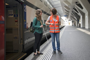 Eine Kundemassistentin der SBB bietet einer sehbehinderte junge Frau den Arm zum Führen an.