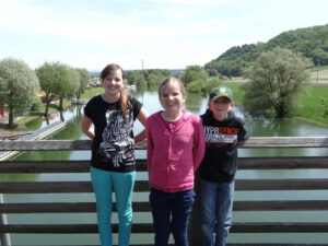 La photo montre trois enfants (de gauche à droite), sur un pont, avec, au second plan, la rivière et la campagne alentour.