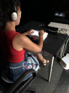 Eine Frau sitzt mit Kopfhörern und Augenbinde ausgestattet vor einem leuchtenden Touch-Pad, das an weitere Geräte angeschlossen ist. 
