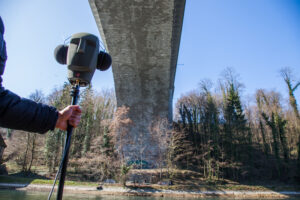 Unter einer Brücke am Fluss hält eine Hand einen Lautsprecher auf einem Ständer, der aussieht wie ein Kopf mit Kopfhörern. 