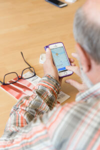Ein Mann sitzt an einem Tisch und hält ein Smartphone in der Hand. Auf dem Bildschirm ist die Karte einer Navigations-App zu sehen. 