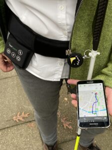 Eine Person mit Langstock und Smartphone in der Hand. Auf dem Smartphone ist eine Navigationsapp zu sehen. 