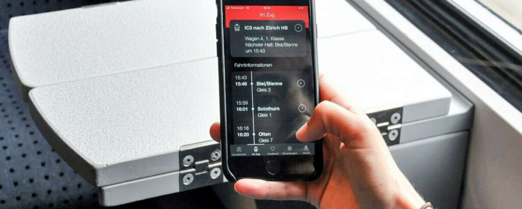 Eine Person sitzt in einem Zug mit einem Smartphone in der Hand. Der Bildschirm ist auf den weissen Schreibmodus auf schwarzem Hintergrund eingestellt.