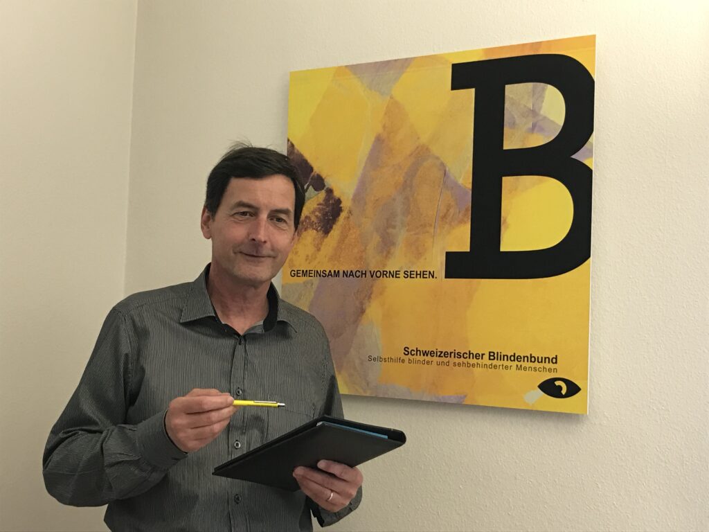 Jvano Del Degan steht vor einem "Schweizerischen Blindenbund"-Plakat, Mappe und Stift in der Hand.