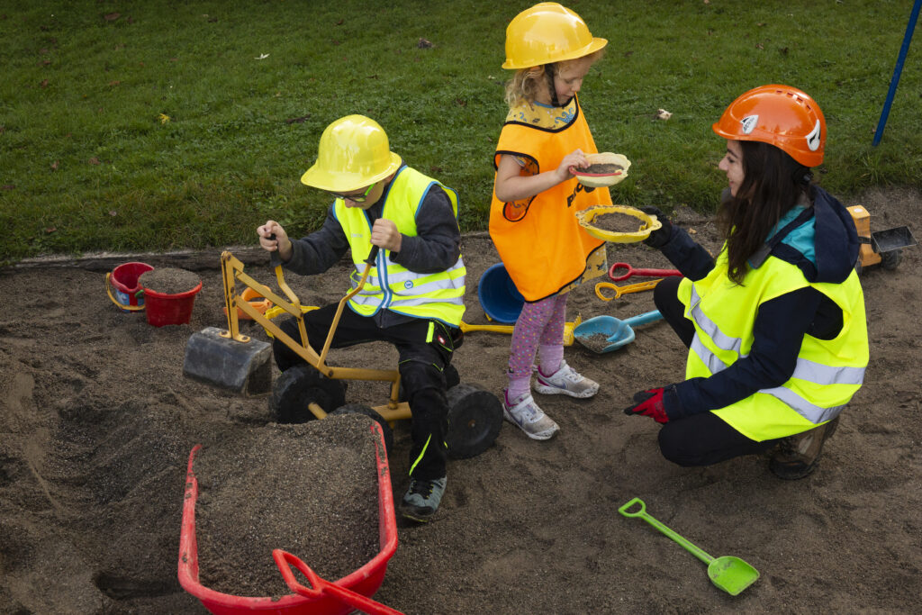 Kinder spielen auf einem Spielplatz in einem Sandkasten. Eine Lehrerin zeigt einem Kind Gegenstände.