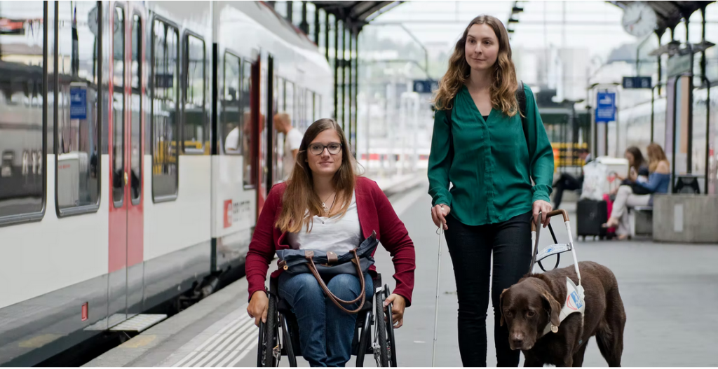 Deux jeunes femmes sur le quai. L'une est assise dans le fauteuil roulant, l'autre marche à côté du fauteuil roulant. D'une main, elle tient la canne blanche, de l'autre, elle tient le harnais de son chien guide.