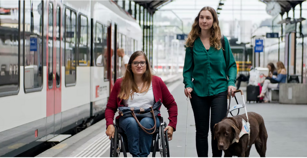 Zwei junge Frauen auf dem Perron. Die eine Person sitzt im Rollstuhl, die andere Person läuft neben dem Rollstuhl. In der einen Hand hält sie den weissen Stock, in der anderen Hand hält sie das Führgeschirr ihres
Führhunds. 