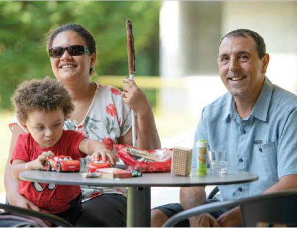 Eine Familie sitzt an einem Gartentisch. Die sehbehinderte Mutter - erkennbar am weissen Stock - hat einen kleinen Jungen auf dem Schoss, der konzentriert mit seinen Autos auf dem Tisch spielt. Vater und Mutter lächeln entspannt in die Kamera.