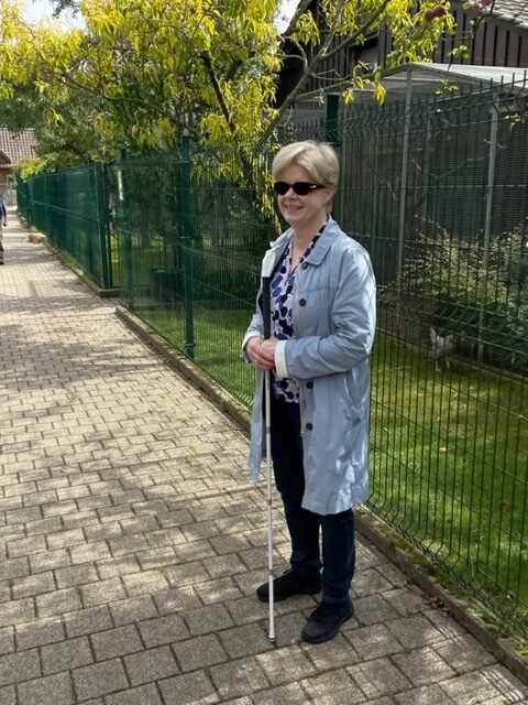 Madame Wagner avec une canne blanche et des lunettes de soleil devant un enclos à poules.