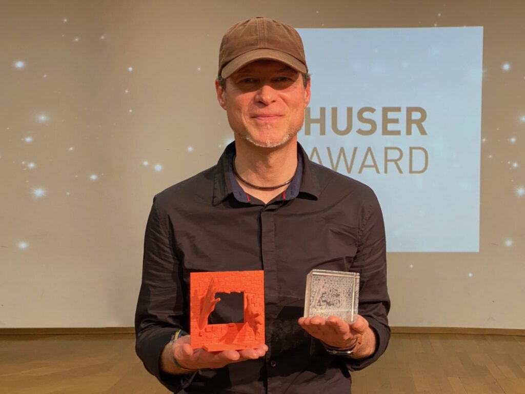 Der Preisträger 2023, Beat Marchetti, hält den Huser Award in Händen und lacht in die Kamera. 