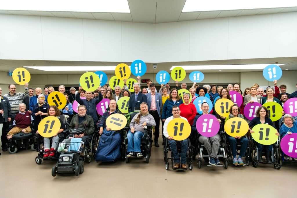 Eine Gruppe von Menschen mit Behinderungen streckt runde Schilder in die Höhe. Auf den Schilder ist ein I mit einem Ausrufezeichen abgebildet.