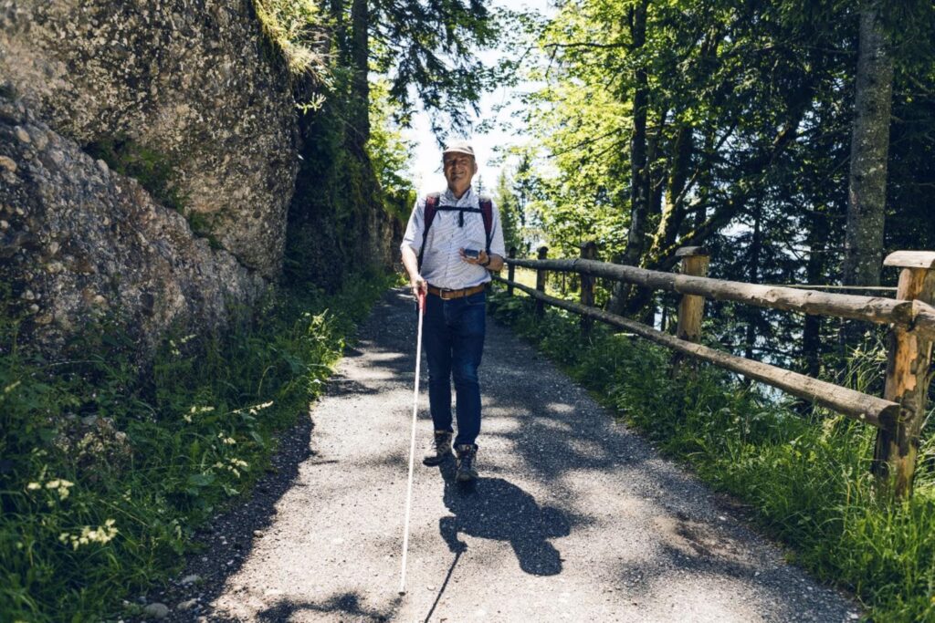 Ein sehbehinderter Mann ist bei strahlendem Wetter auf einem idyllischen Bergwanderweg am wandern.