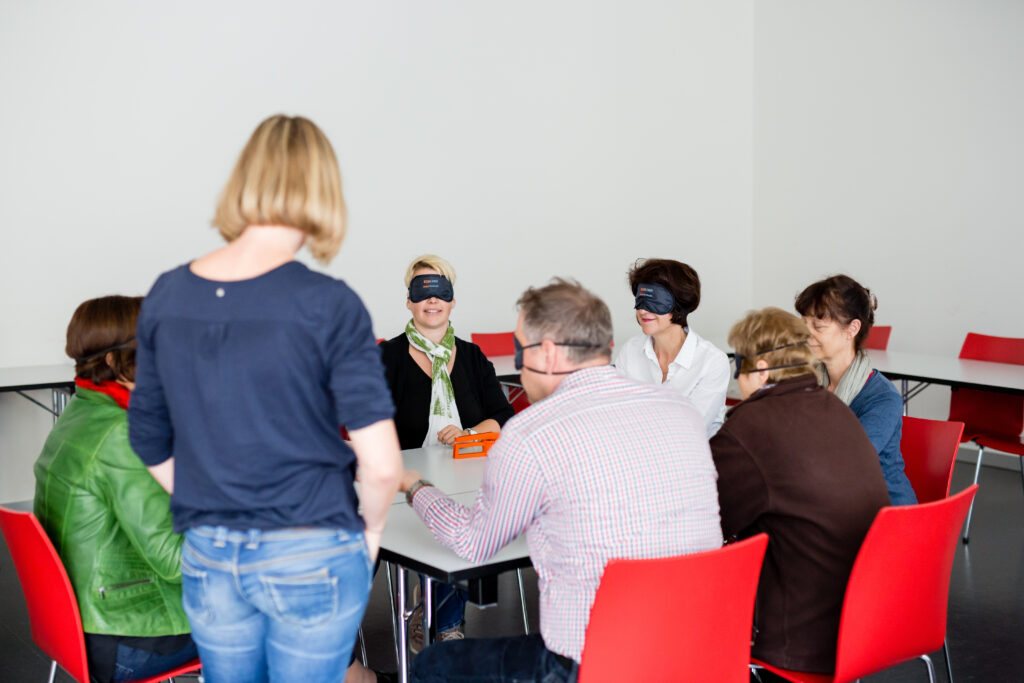 das Bild zeigt eine Gruppe Menschen, die mit Dunkelbrillen Aufgaben lösen. 