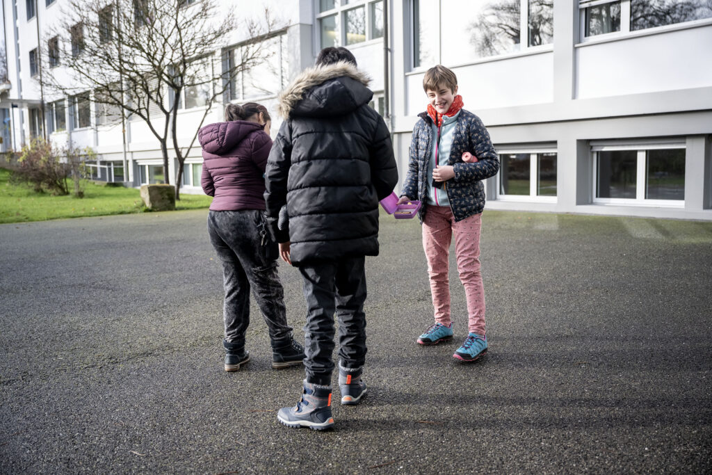 drei Kinder stehen in einer Gruppe auf einem Schulhausplatz.