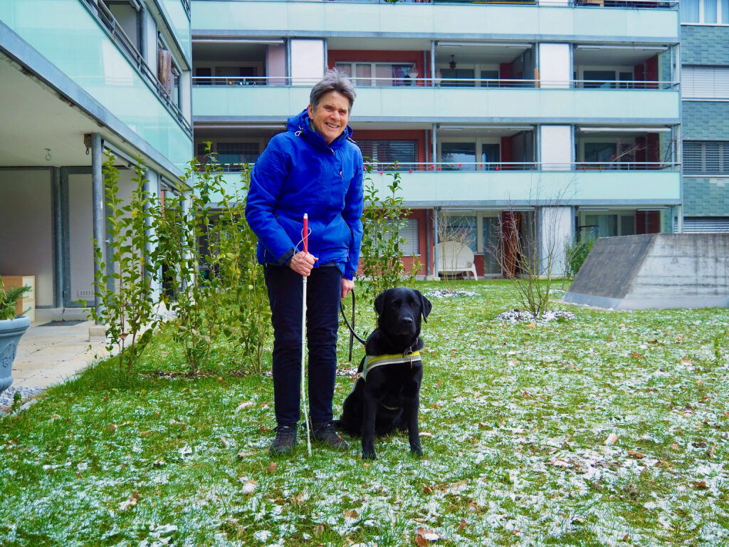 Maria Theresia Müller steht mit ihrer Labradorhündin Henni vor einer Häuserzeile im Garten. 