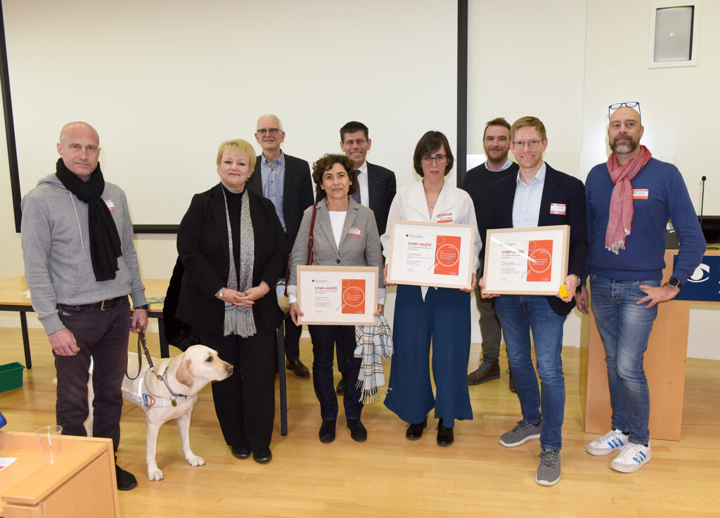 Photo de groupe avec, au centre, notamment les lauréats du prix récompensant des offres de formations accessibles. Se partagent le premier prix : le projet « Taktile Fokuswörter zur Unterstützten Kommunikation » de la Haute école intercantonale de pédagogie curative HfH de Zurich et du Sonnenberg, la bibliothèque MonaLira de l’Association Plein Accès et le musée « Voir autrement » à Zollikofen. Les lauréats tiennent un document en main.