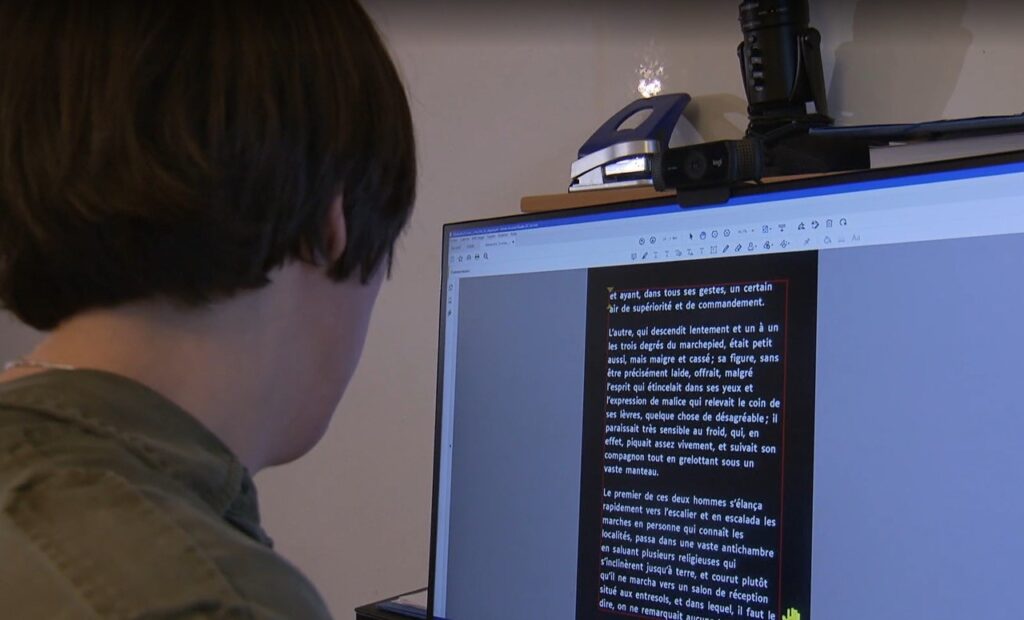 Blick über die Schulter einer Person, die vor dem Computer sitzt. Auf dem Bildschirm ist ein französischer Text in weisser Schrift und vergrösserten Buchstaben dargestellt.