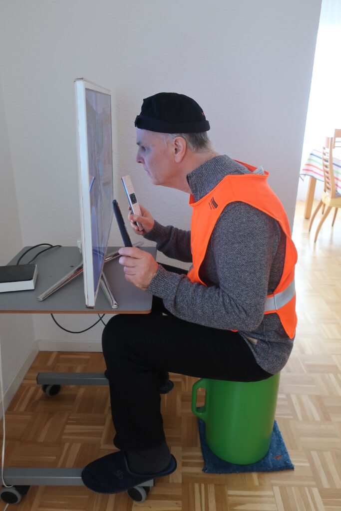 Un homme est assis à une table devant un téléviseur. Il tient sa tête très près de l’écran et utilise deux télécommandes, une dans chaque main, pour manipuler l’appareil.