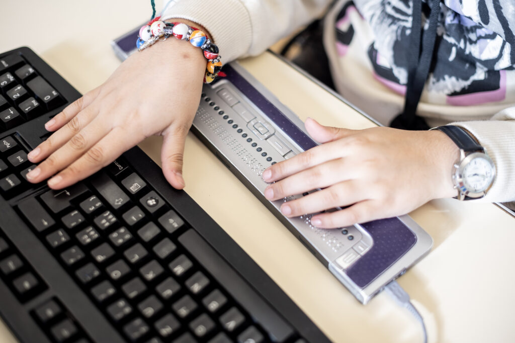 Deux petites mains utilisent un afficheur braille placé devant un clavier d’ordinateur. La main gauche repose sur le clavier, tandis que la main droite lit les points en braille.