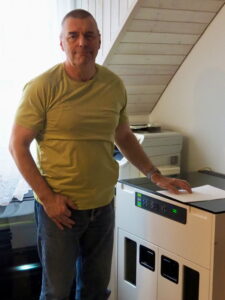 Un homme se tient à côté d’une imprimante braille et tient une feuille de papier à la main. Il regarde en direction de la caméra. À l’arrière-plan, une autre imprimante est visible.