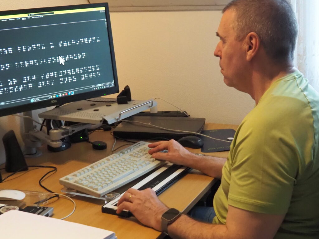 Un homme est assis à un bureau et travaille sur un ordinateur. L’écran affiche des caractères en braille. Devant le clavier se trouve une plage braille, que l’homme lit de la main gauche