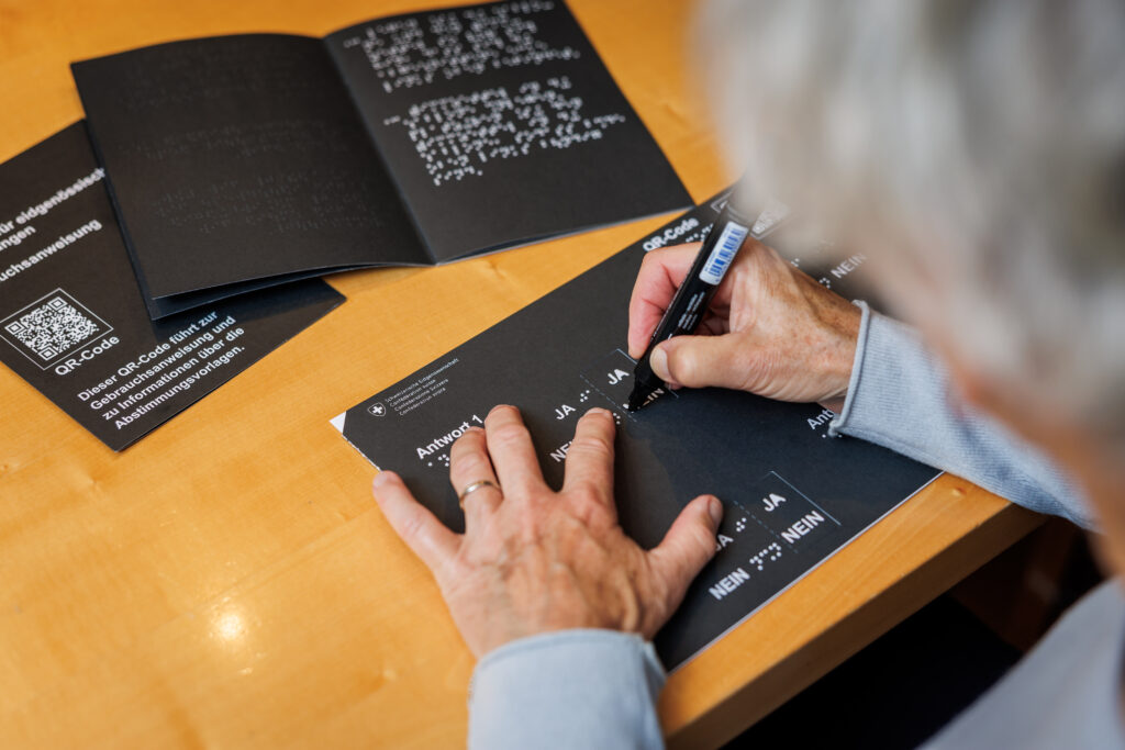 Sont éparpillées sur une table les instructions en braille et en noir concernant le modèle de vote tactile qu’une dame est en train d’utiliser.