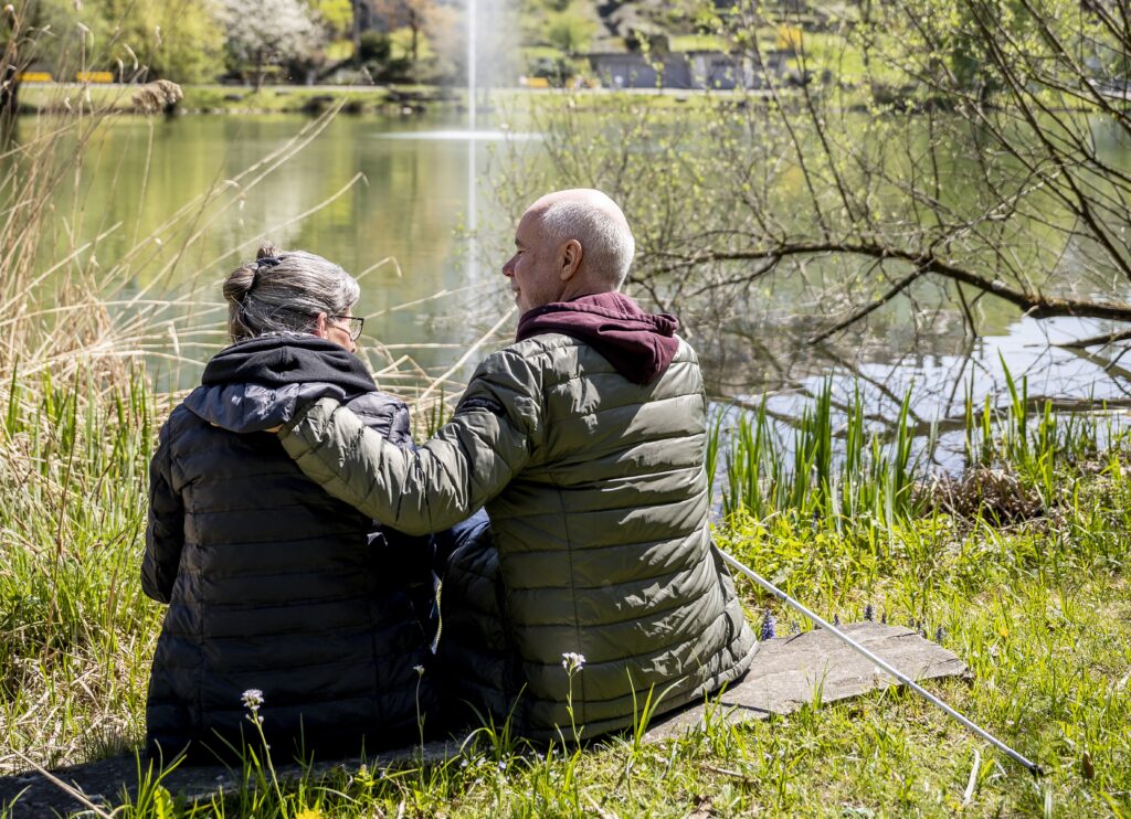 Vus de dos, une femme et un homme sont assis, vêtus de vestes d’hiver, au bord d’un étang. L’homme entoure de son bras gauche les épaules de la femme.