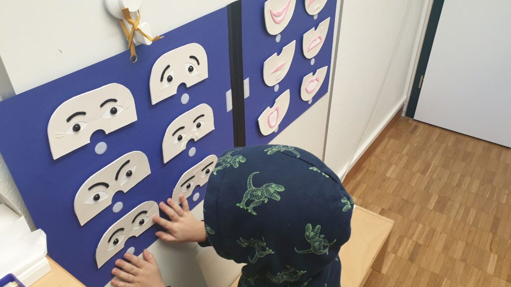Un poster bleu avec des masques tactiles montrant différentes expressions faciales est accroché au mur. Un enfant tâte les masques.