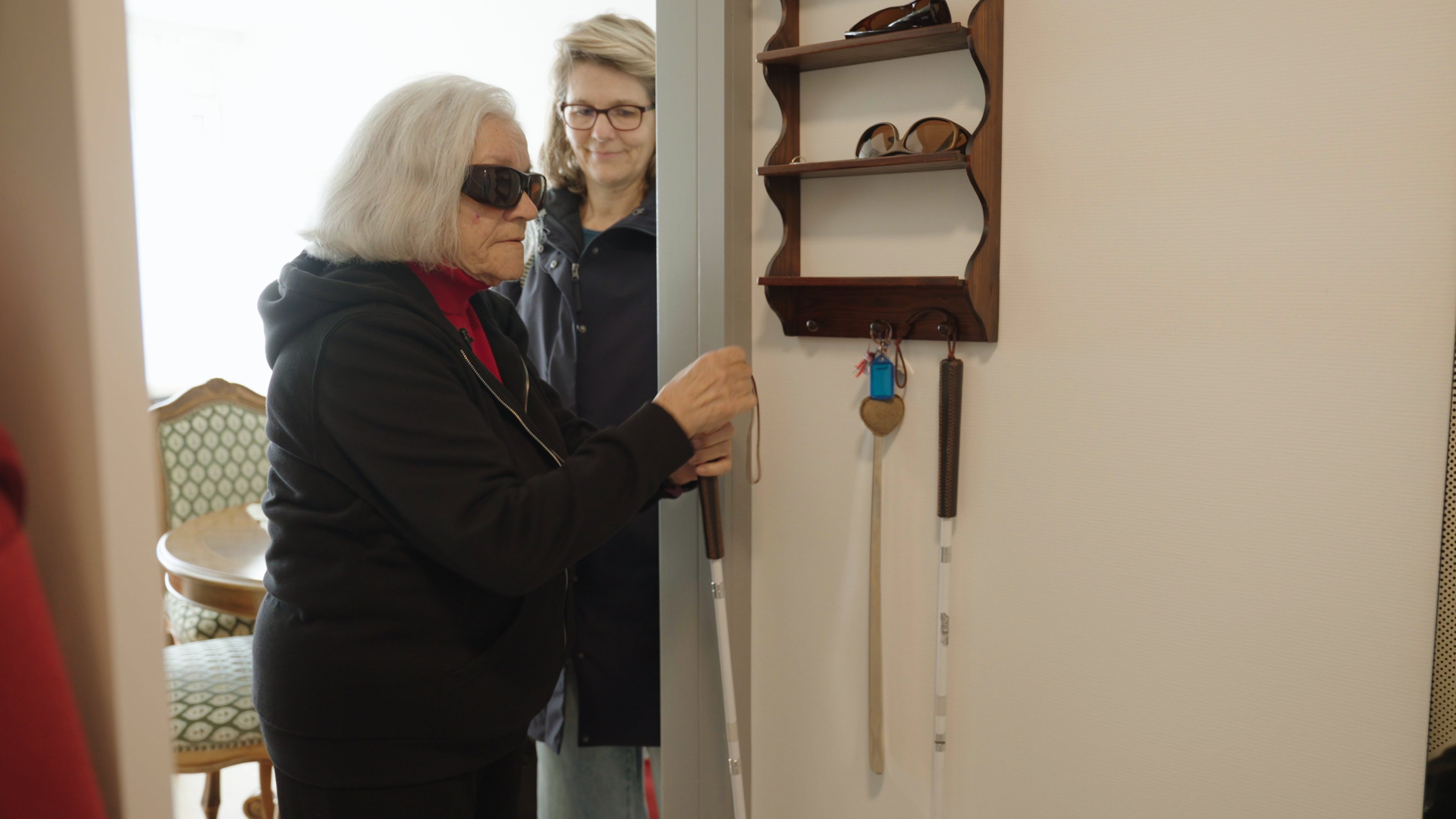 Eine ältere Frau mit Sonnenbrille steht im Flur vor einer Garderobe. Sie hat gerade ihren weissen Stock vom Haken genommen. Im Hintergrund ist eine andere Frau zu sehen.