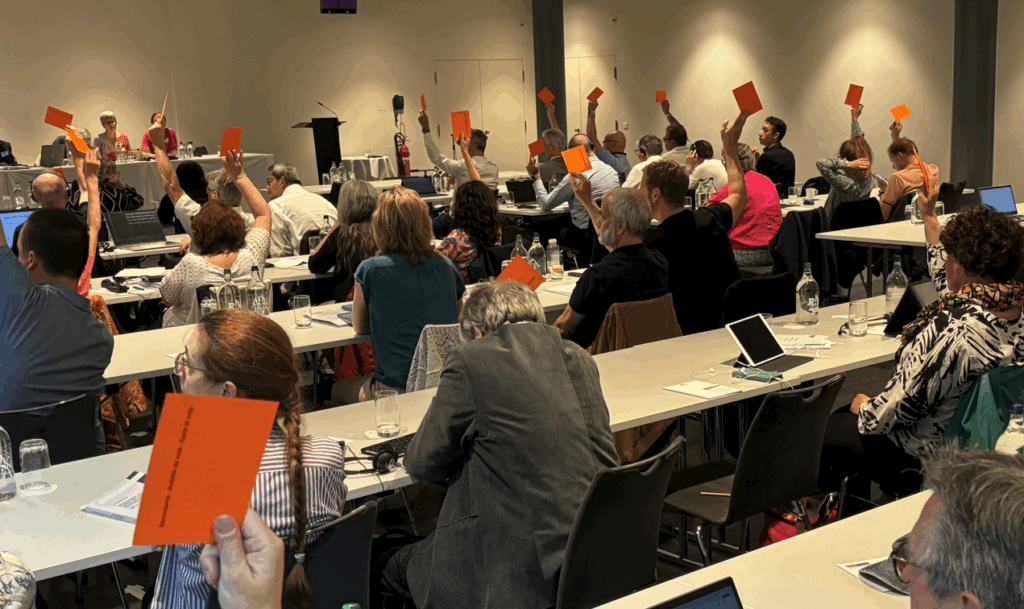 In einem Raum stehen mehrere Tischreihen, an denen rund dreissig Personen sitzen. Einige von ihnen heben einen orangefarbenen Zettel in die Höhe.