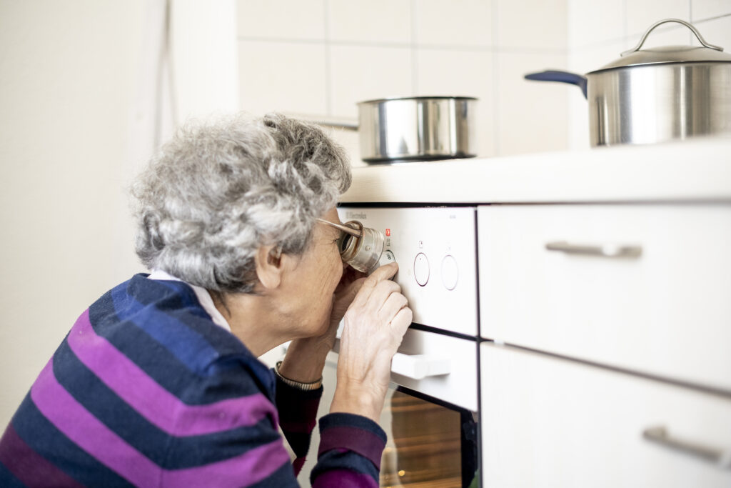 Eine ältere Frau mit Brille und befestigtem Vergrösserungsglas beugt sich dicht an den Drehknopf eines Bachkofens, um die Temperatur abzulesen. 
