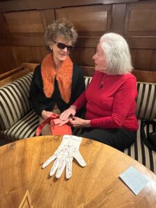 Zwei ältere Frauen sitzen auf einer Bank. Vor ihnen steht ein Tisch, auf dem ein Handschuh mit Lormzeichen liegt. Die Frau auf der rechten Seite berührt mit ihrer rechten Hand die Handinnenseite der linken Hand der anderen Frau.