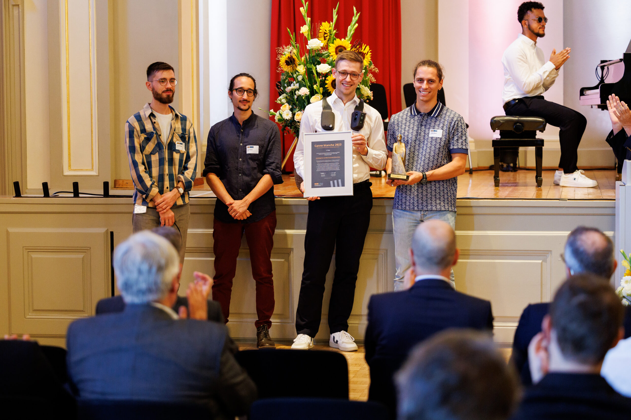 Alternativtext: Drei junge Männer stehen auf einer Bühne. Hinter ihnen befindet sich ein grosses Blumengesteck. Der Mann in der Mitte trägt ein Gerät über den Schultern und hält eine Auszeichnung in der Hand. Der Mann neben ihm hält die Gewinnerstatue des Prix de la Canne blanche in der Hand.