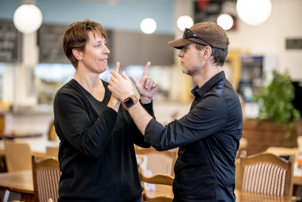 Debout face à face dans un café, un homme et une femme communiquent en langue des signes tactile. A gauche, la femme forme des lettres avec ses mains. A droite, l’homme touche ses mains pour appréhender les signes. Vêtu d’une chemise foncée, il porte une casquette et des lunettes noires sur la tête